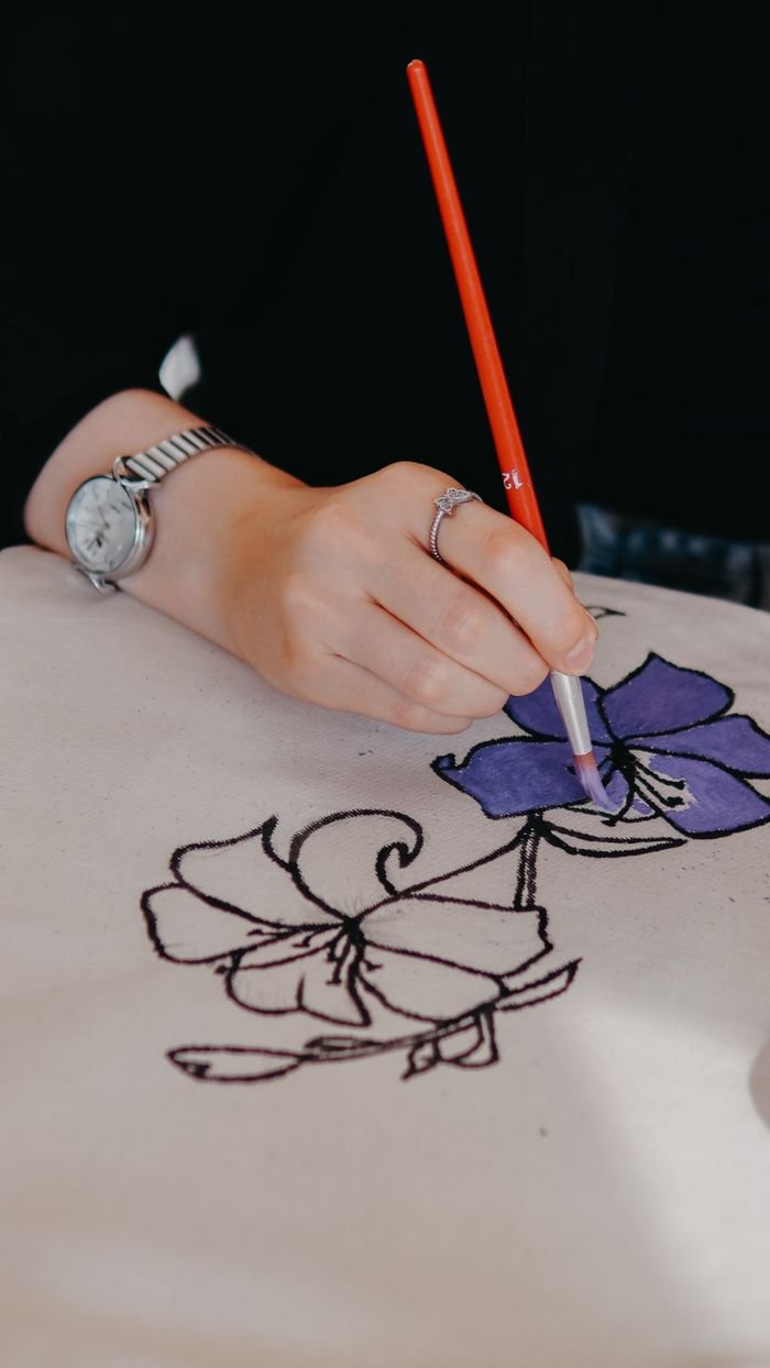 Hand paints intricate floral design on fabric with red-handled brush, showcasing creativity.