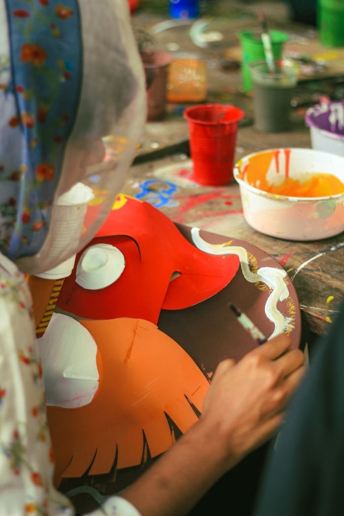 A South Asian student paints a vibrant, traditional mask in Dhaka classroom setting.